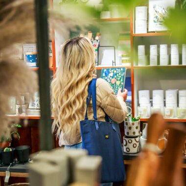 A woman with blond hair shops in a home decor store, holding a notebook and wearing a blue bag.